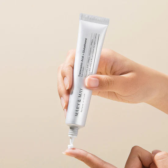 Hand holding a tube of Mary & May Tranexamic Acid + Glutathione Eye Cream 30ml skincare product against a beige background